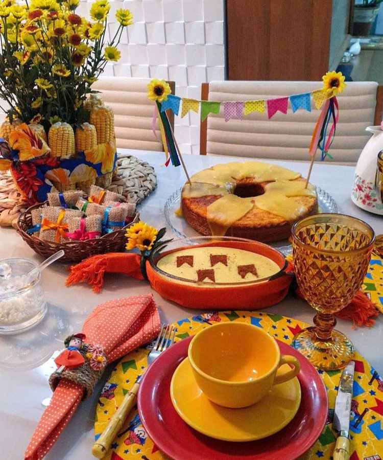 A imagem representa uma decoração de festa junina colorida. Contém mesa com comidas como bolo e cural, além de enfeites coo bandeirinha, milho, flores, etc