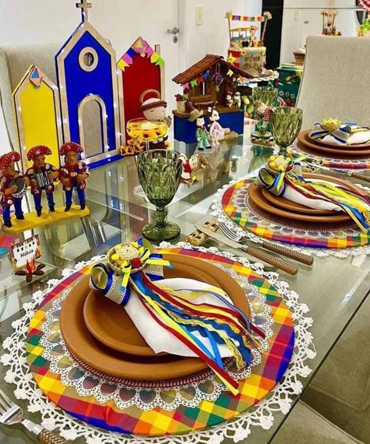 A imagem representa decoração para festa junina. Contém pratos, sousplats coloridos, guardanapos enfeitados com fitas de cetim, laços e mini-chapéu de palha, além de bonecos típicos