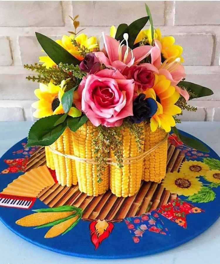 A imagem representa uma decoração de festa junina. Contém arranjo de flor enfeitado com espigas de milho, sobre souplat de festa junina