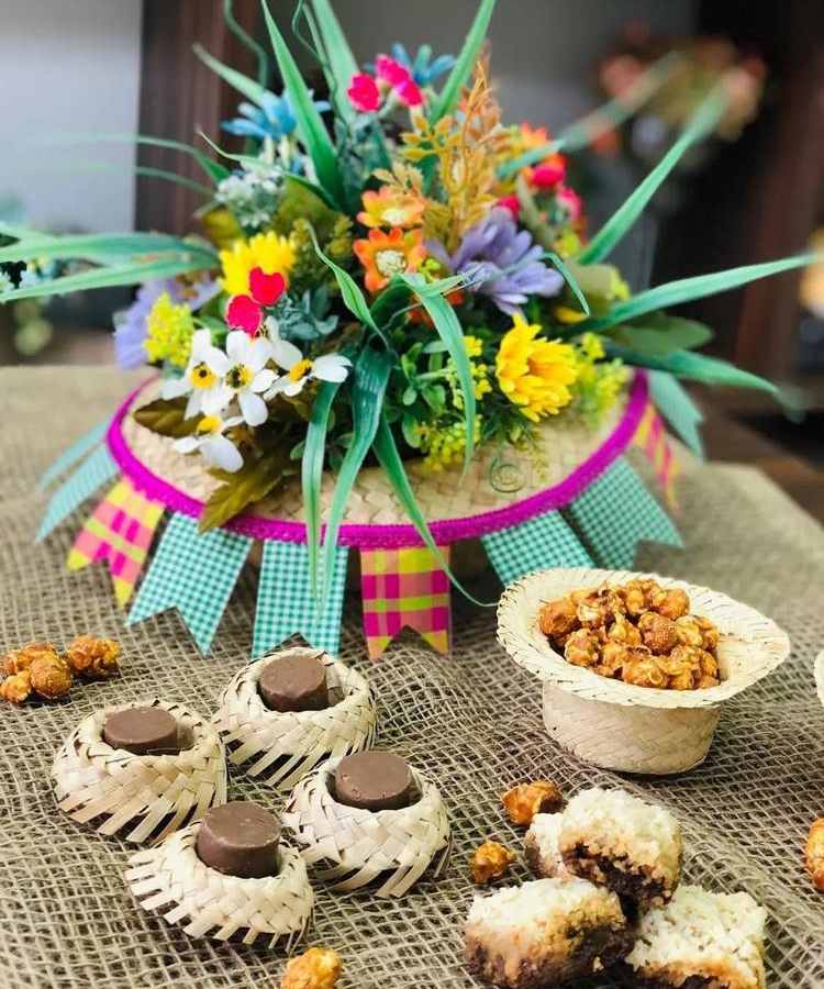 A imagem representa uma decoração de festa junina colorida. Contém xapéu decorado com bandeirinhas e flores dentro, além de mini chapéus com doces de chocolate e pipoca
