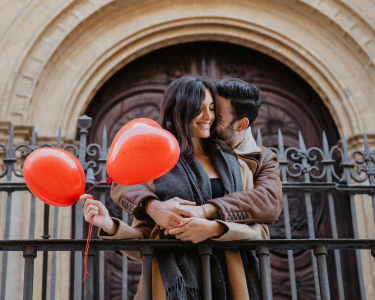 Homem abraçando mulher, que está segurando balões de coração, perto de portão de ferro: dia dos namorados