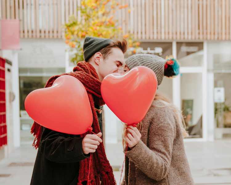 Casal se beijando e segurando balões de coração vermelho: dia dos namorados