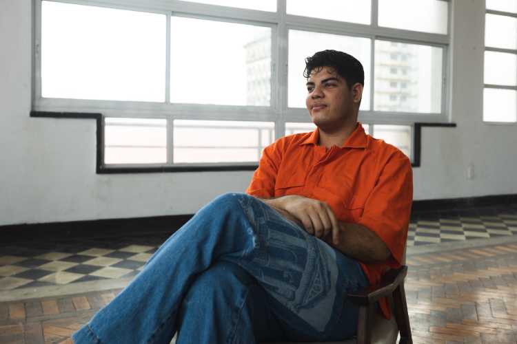 Levi Santos sentado em cadeira, veste camisa laranja e calça jeans