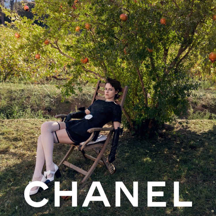 Atriz Kristen Stewart sentada em uma cadeira, em frente uma árvore, usando um vestido preto, luvas, meias longas e um par de sapatos Mary Jane preto e brancos da Chanel.