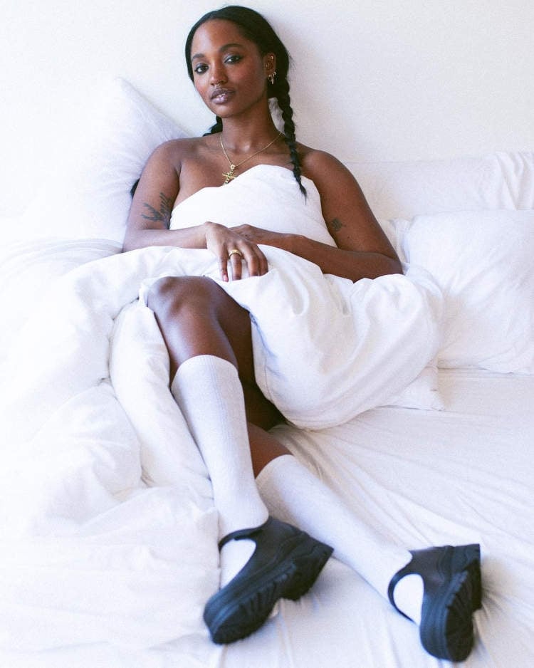 Mulher deitada em uma cama com fundo branco. Ela está usando meias brancas de cano longo e sapatos Mary Jane pretos.