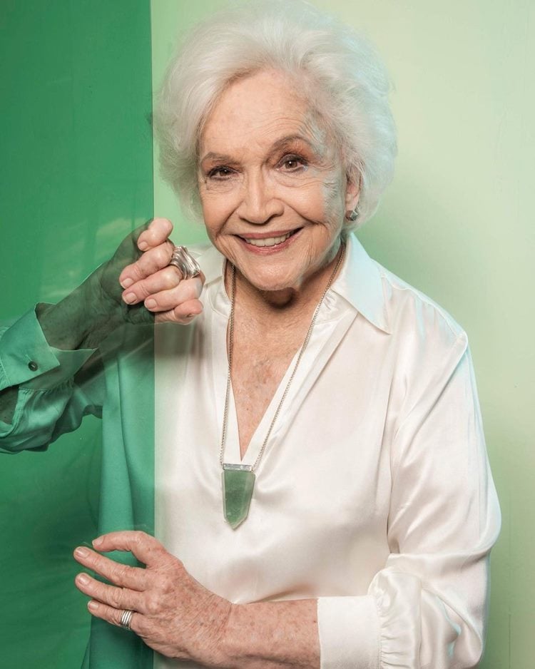 Atriz em ensaio de 2019 em comemoração aos seus 90 anos. Ela segura porta de vidro verde, usa camisa de seda e colar com pedra verde.