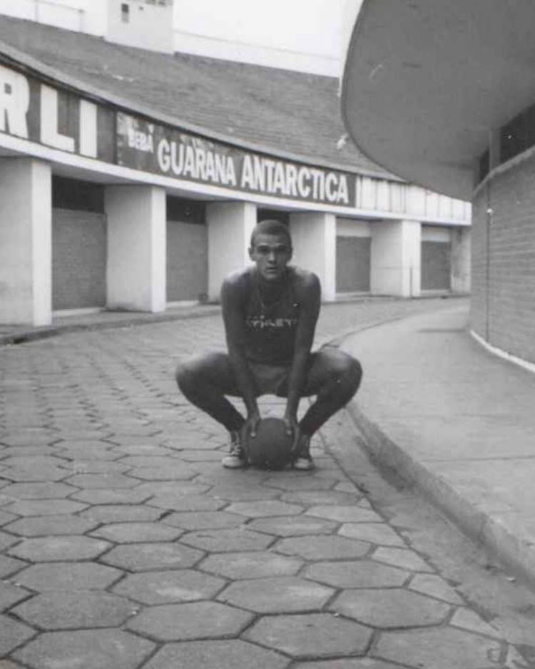 Oscar Schmidt agachado com bola na mão sentado em rua com lojas vendendo guaraná antarctica, foto antiga em preto e branco.