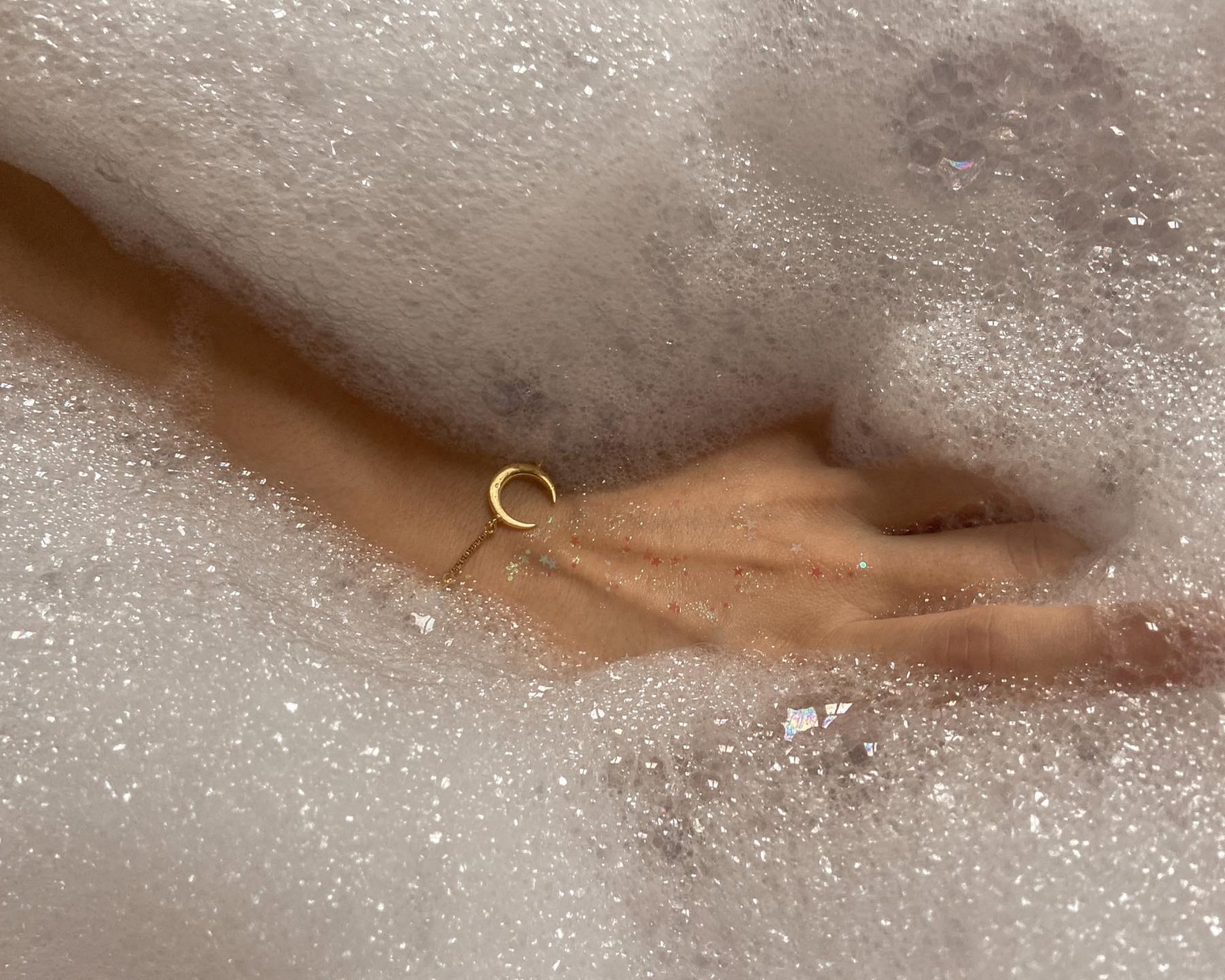 mão feminina com pulseira de ouro em formato de uma lua crescente em uma banheira de espuma