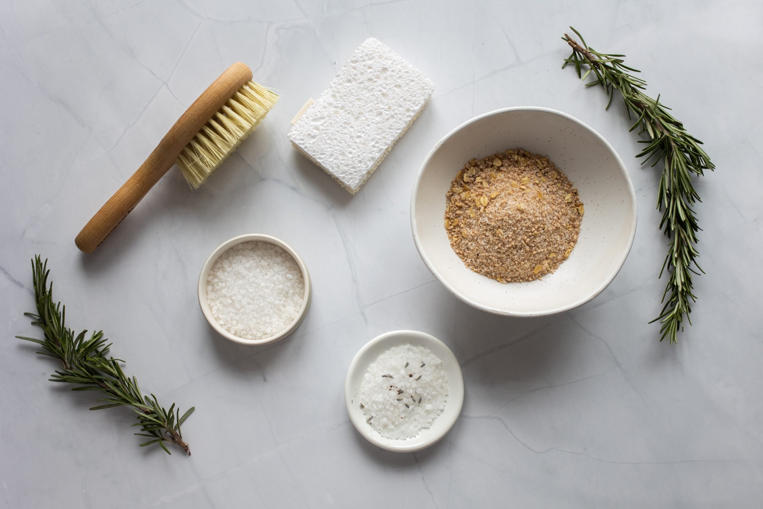 bucha e escova de banho com potes de sal e açúcar e ramos de alecrim espalhados em uma superfície branca