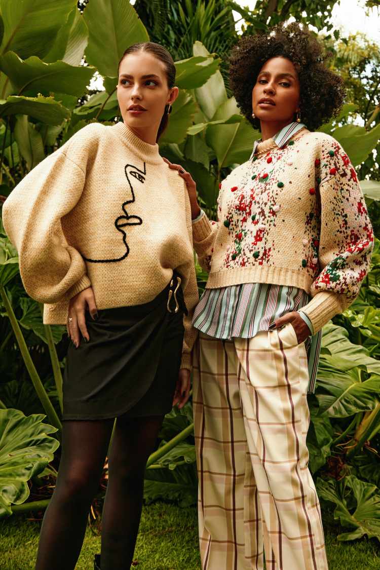 duas mulheres em funfo de plata, a esquerda veste tricô bege e saia com meia calça pretas. A direita, tricô bege e calça de mesma cor xadrez