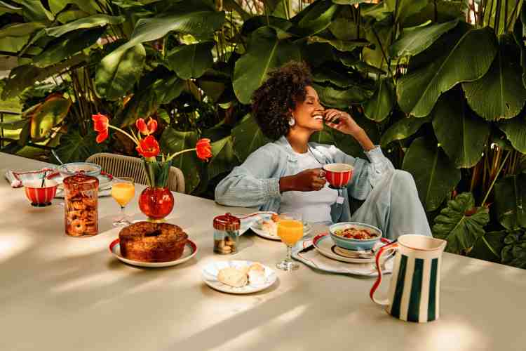 mulher sentada com mesa de café da manhã, segura xícara em fundo de plantas