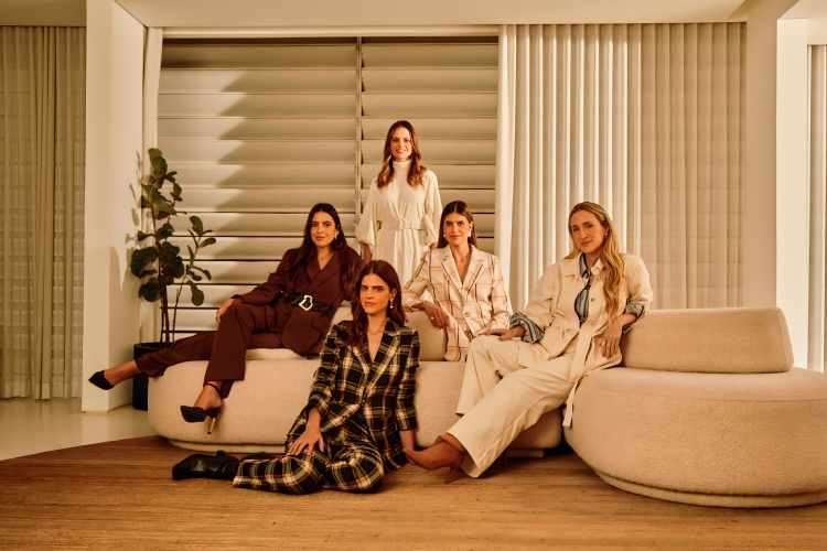 cinco mulheres sentadas e apoiadas em sofás em ambiente claro, todas vestem peças de alfaiataria da coleção Gallerist para Riachuelo