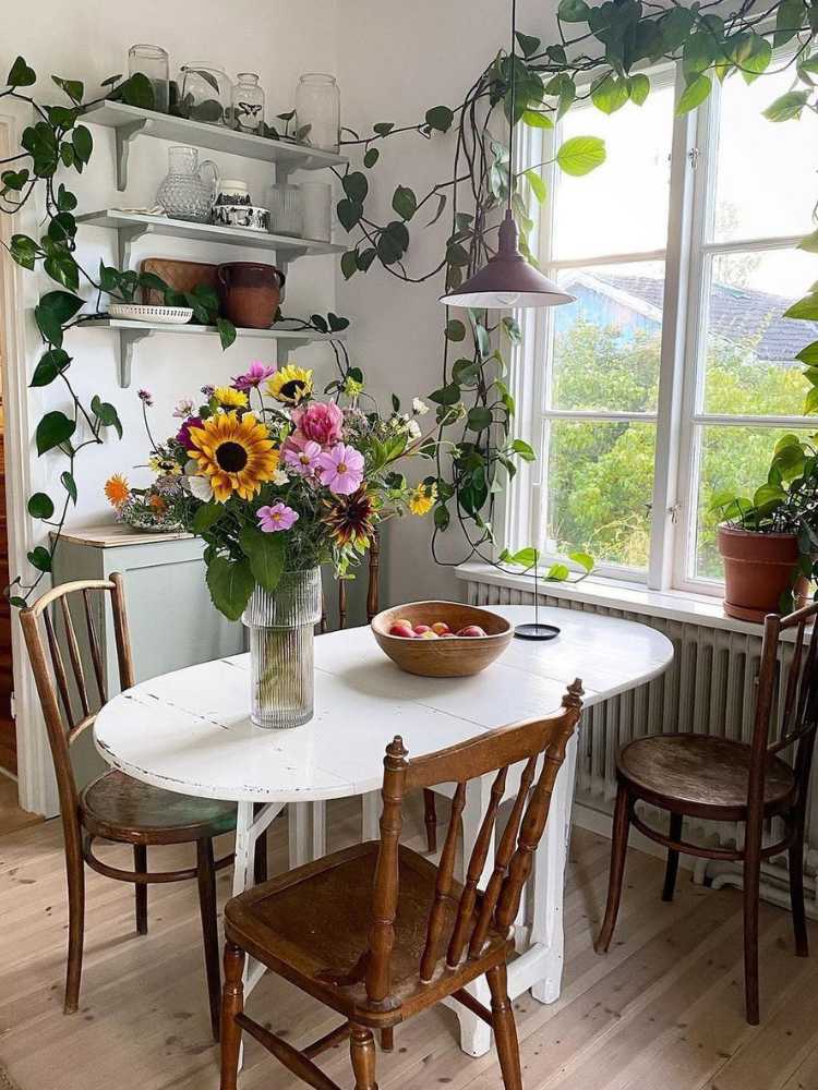 Copa com mesa branca, cadeira de madeira, janela com vista para montanha e diversas plantas na decoração, pendentes e em vasos 