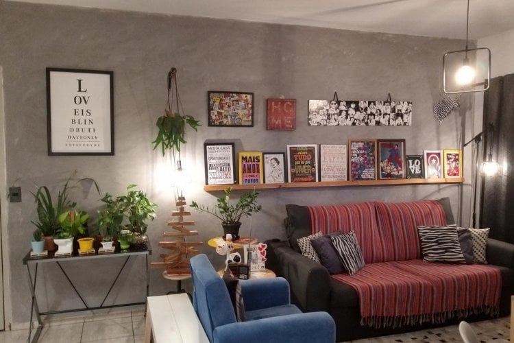 Foto de sala com parede de cimento queimado, quadros na parede, sofá azul e cinza, almofadas e plantas no canto, em aparador e penduradas