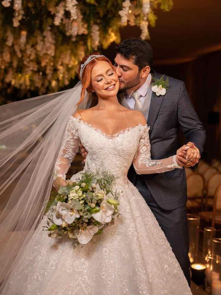 Mirela Janis, do Power Couple, de noiva e seu noivo a beijando no rosto em seus casamentos