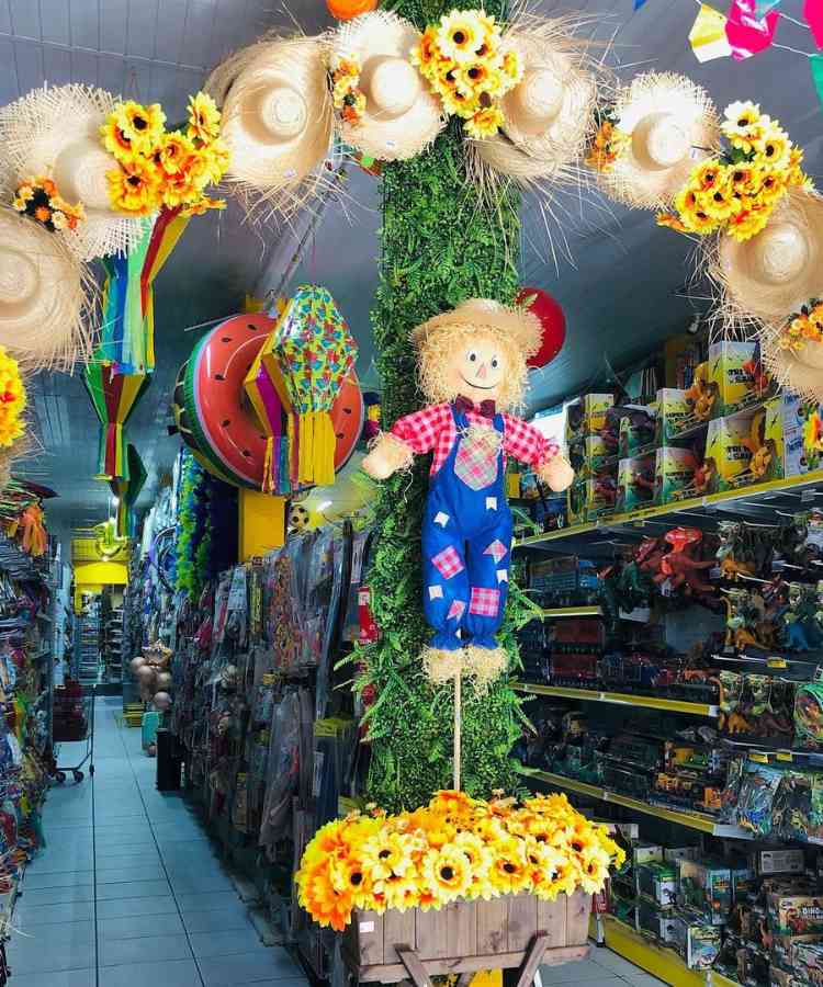 Ideia de decoração de vitrini com arco de chapéus de palha e girassóis, boneco espantalho, balões e caixote com flores de girassol