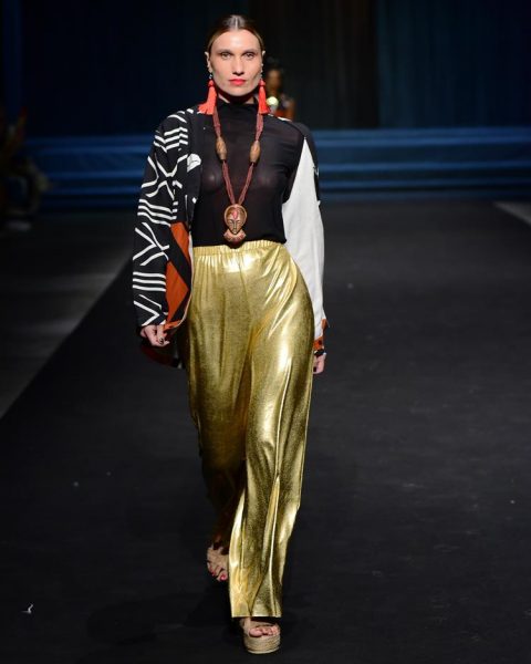 modelo no desfile Liana D'Áfrika com calça metalizada dourada, blusa preta e casaco estampado com laranja.