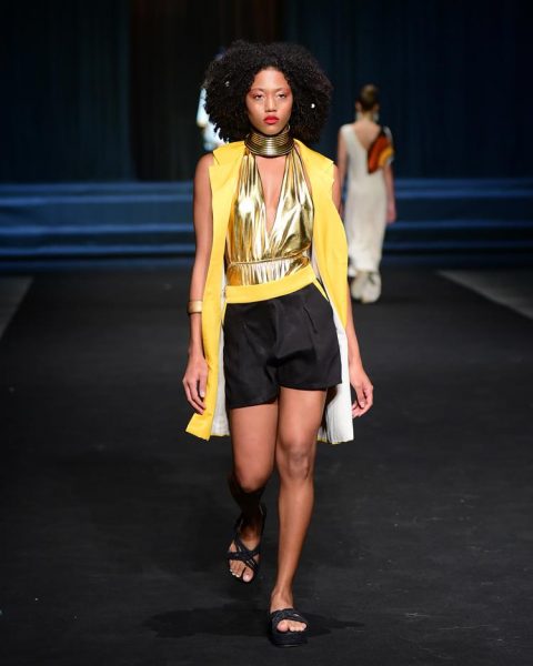 modelo no desfile Liana D'Áfrika com shorts preto, cinto e colete amarelos e body metalizado dourado.