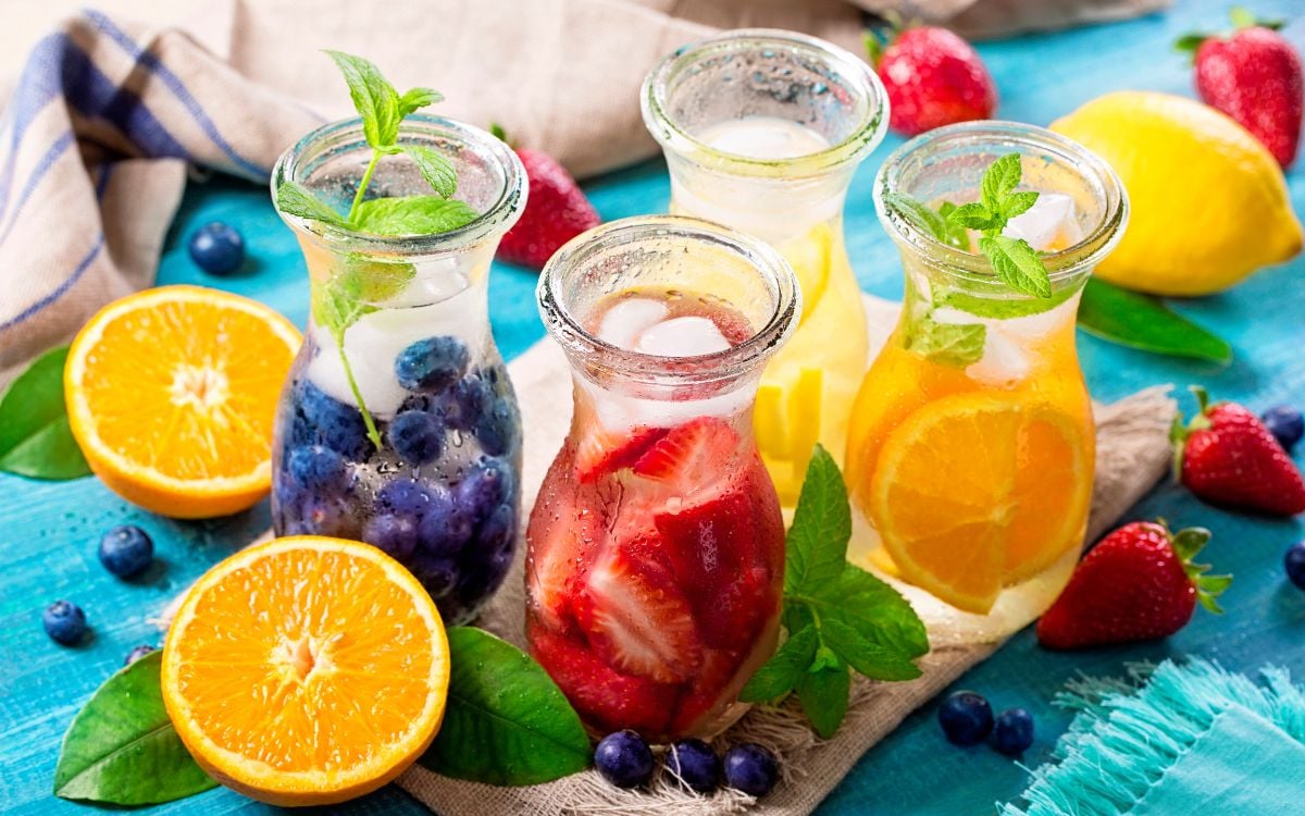 Quatro copos com águas saborizadas, com frutas picadas morango, laranja, limão e bluberry, em tábua de madeira, com as mesmas frutas sobre ela