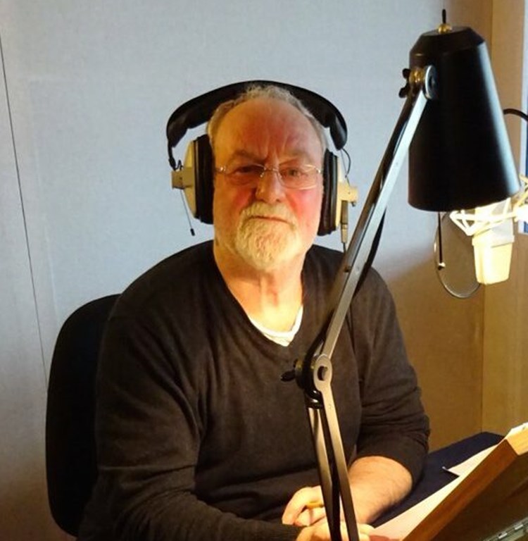 Ator Bernard Hill com fone de ouvido em cadeira com mesa e luminária