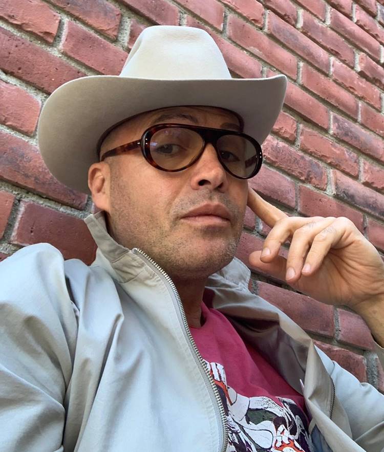 Ator Billy Zane, de Titanic, hoje em dia. Encostado em parede de tijolinhos, usando chapéu, óculos, camiseta e jaqueta