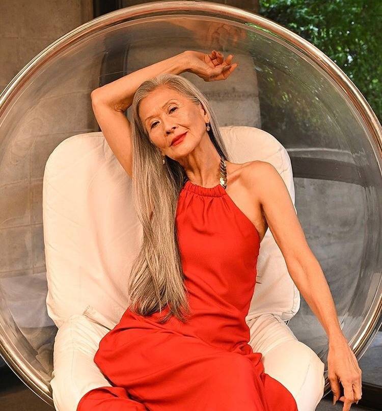 Foto de modelo Rosa Saito, que possui fios naturais longos grisalhos, usando vestido vermelho sentada em balanço.