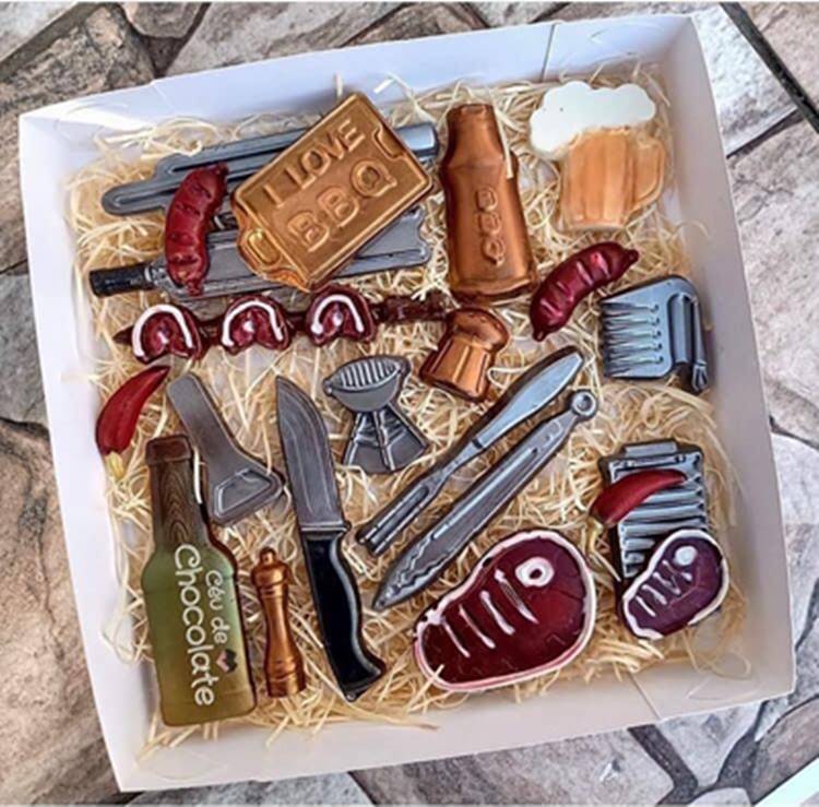 Uma cesta para pais amantes de chocolate, com itens como uma faca, carne, churrasco e cerveja, todos feitos de chocolate em um trabalho artesanal.