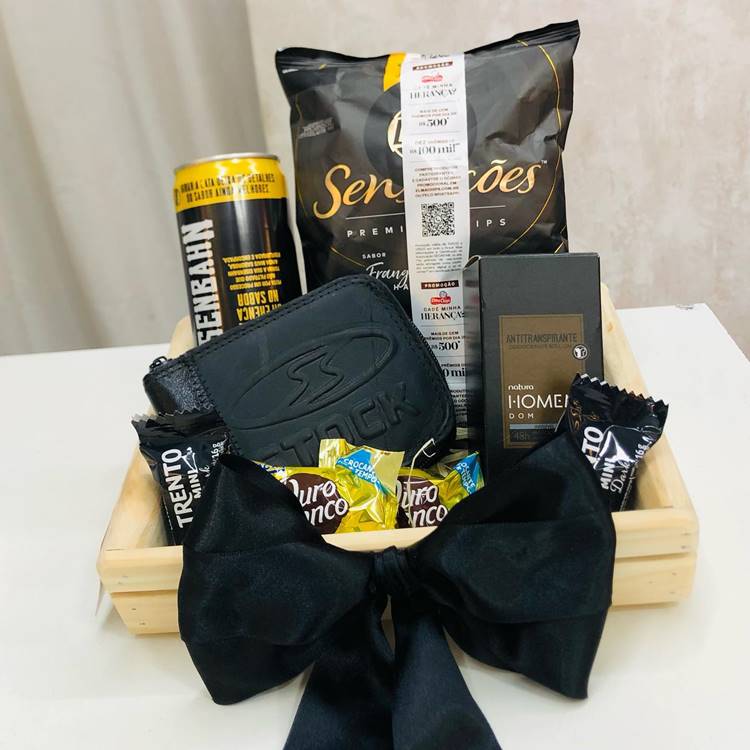 Uma cesta preta contendo salgadinhos, cervejas, carteira, chocolates e perfume, formando uma montagem.
