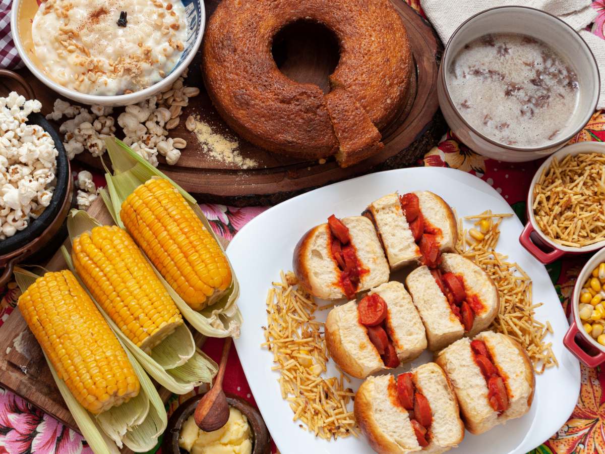 Mesa com comidas típicas de festa junina, como milho cozinho, cachorro quente, canjica, bolo de fubá, etc