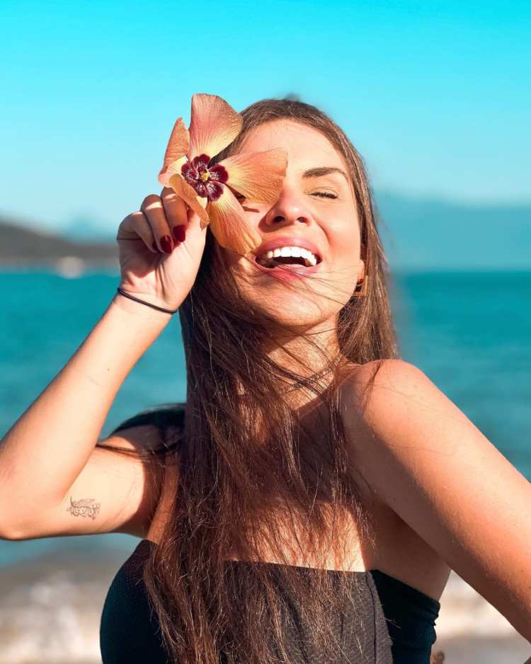 mulher posando segurando uma flor no rosto, ao fundo praia