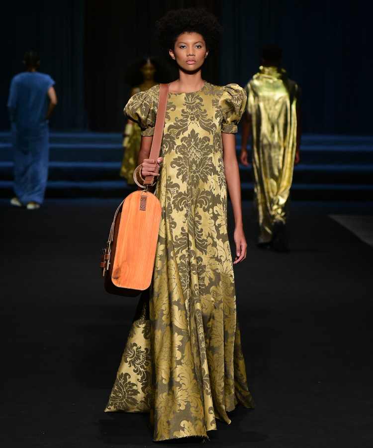 Modelo usando vestido come stampa floral dourado e bolsa de madeira no desfile de Linbebergue no DFB Festival 2023