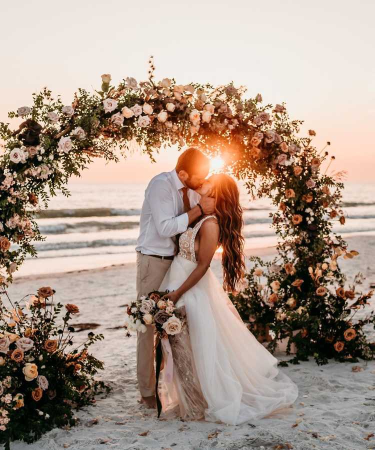Noivos se beijando em arco de flores em casamento na praia