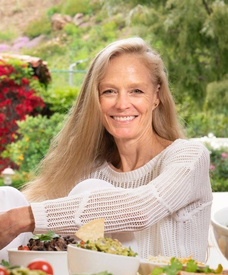 Atriz Suzi Amis hoje em dia, em 2023, sentada em mesa com vários potes de comida