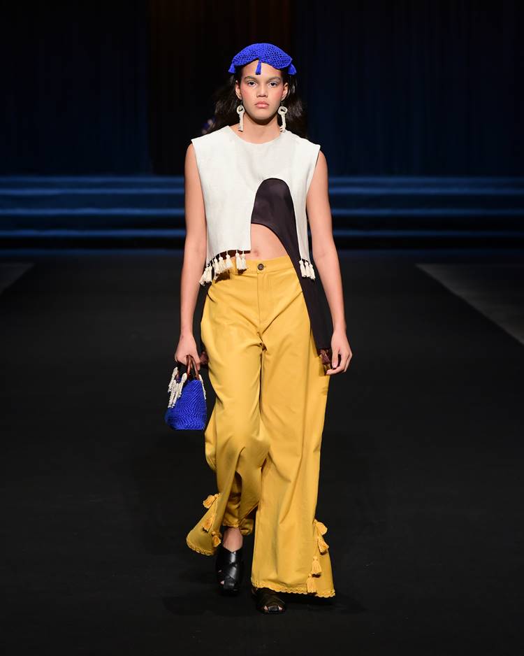Modelo com blusa off-white + marrom + calça amarela + bolsa azul + acessório de cabelo azul em crochê no desfile da UDESC no Dragon Fashion Brasil 2023
