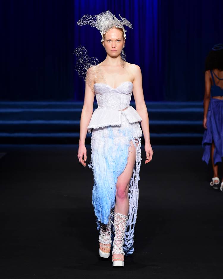 Modelo com cropped branco + saia branca e azul + acessório de cabeça transparente no desfile da UNIFOR no Dragon Fashion Brasil 2023