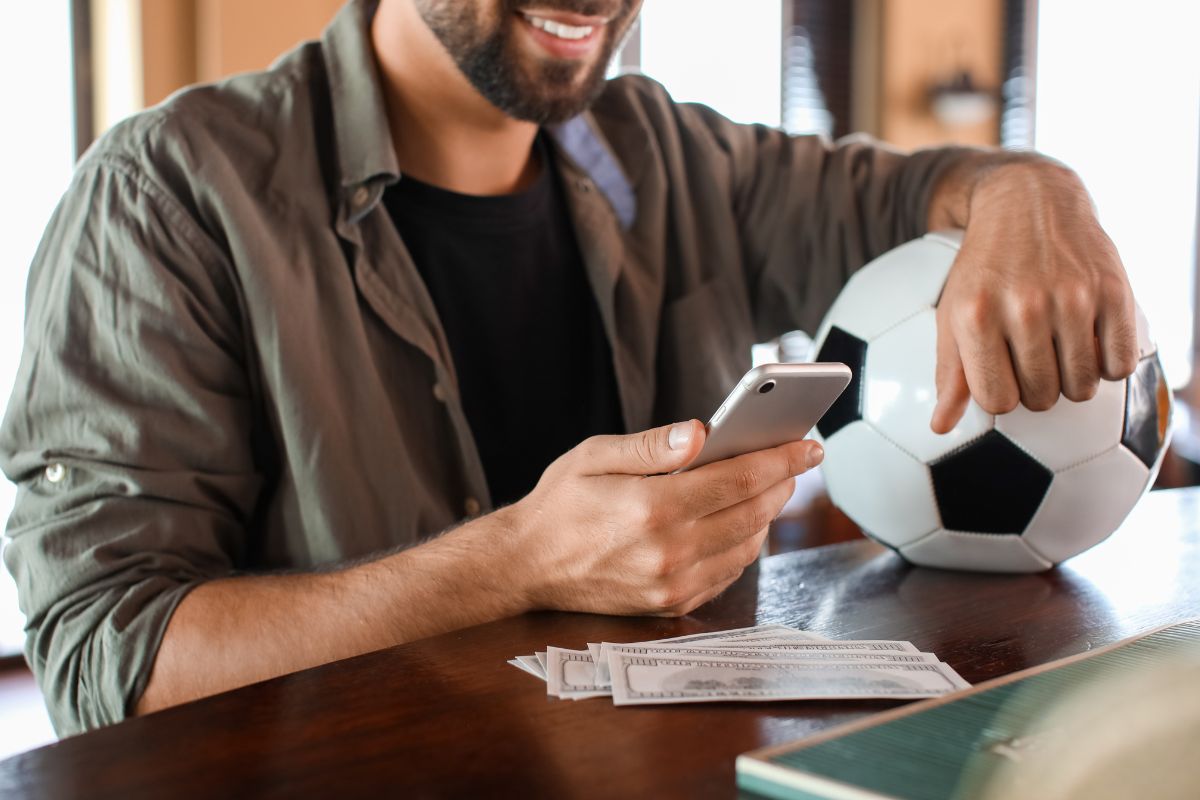 homem segurando bola de futebol e celular enquanto faz apostas esportivas online