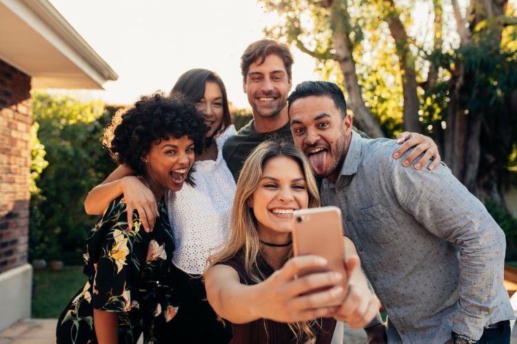 Amigos tirando selfie, envolvendo duas mulheres de pele clara, uma mulher de pele negra e dois homens de pele clara