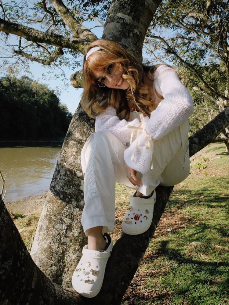 mulher sentada em uma árvore usando tricô, calça cargo e crocs, tudo em off white