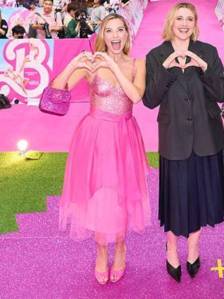 Looks Margot Robbie Barbie "Day-to-night" de 1985: atriz usa vestido pink, faz coração com as mãos e está acompanhada de outra atriz