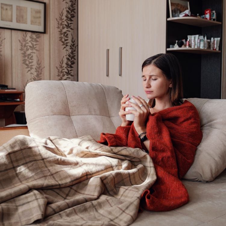 mulher sentada em sofá coberta e com xícara na mão