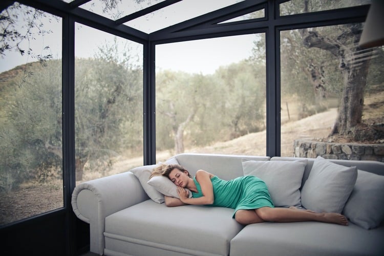 Mulher com um vestido verde dormindo no sofá de uma casa com grandes janelas de vidro com vista para a natureza 