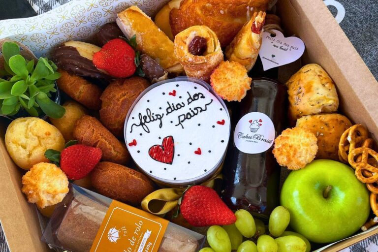 A imagem mostra uma cesta de Dia dos Pais repleta de itens comestíveis variados, como frutas frescas, pães, doces e uma garrafa de bebida. Há um cartão com texto e um biscoito decorado com a frase “Feliz dia dos Pais”. A diversidade e apresentação tornam a cesta visualmente atraente.