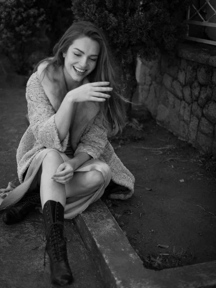 Mulher não-modelo com mais de 30 anos em ensaio fotográfico, usando casaco e botas, sentada em paralelepípedo 