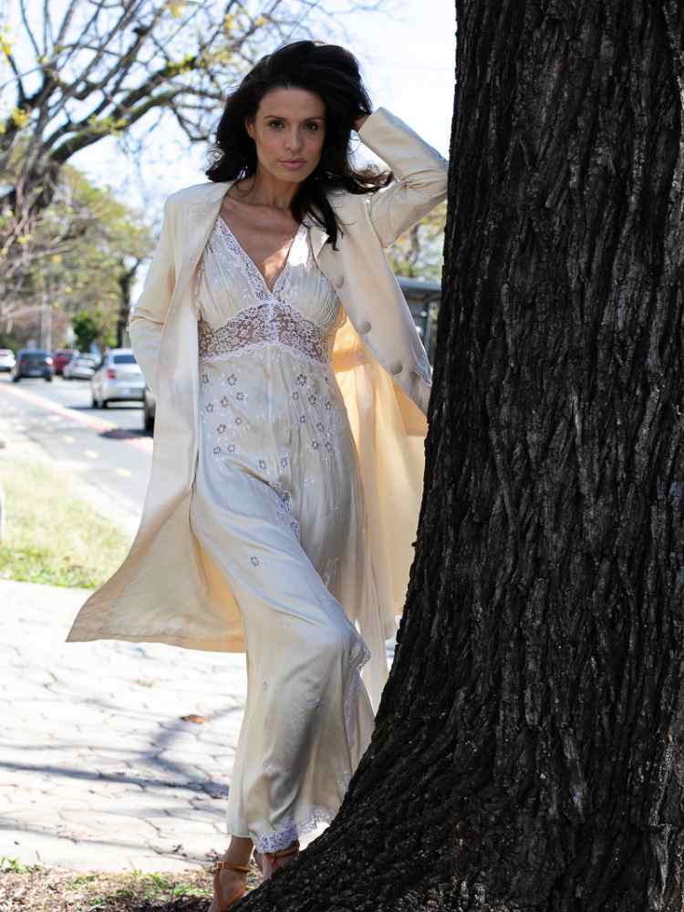 Mulher não-modelo com mais de 50 anos em ensaio fotográfico, usando vestido de renda + blazer, encostada em árvore de rua 