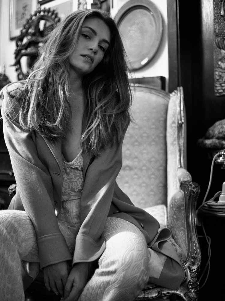 Mulher não-modelo com mais de 30 anos em ensaio fotográfico, usando body de renda, calça texturizada, blazer, sentada em poltrona. Foto preto e branco