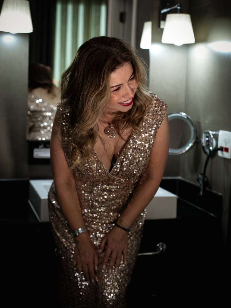 Mulher não-modelo com mais de 40 anos em ensaio fotográfico, usando vestido de paetês dourado, em banheiro, sorrindo