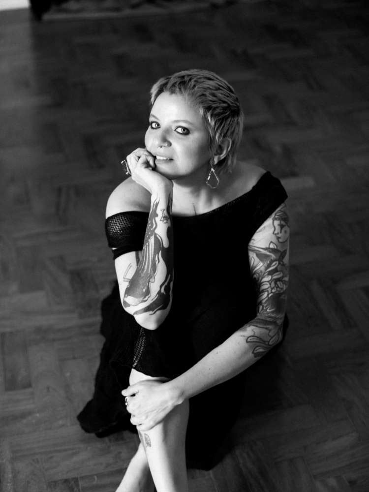 Mulher não-modelo com mais de 40 anos em ensaio fotográfico, usando vestido preto, sentada em chão de madeira. Foto preto e branco