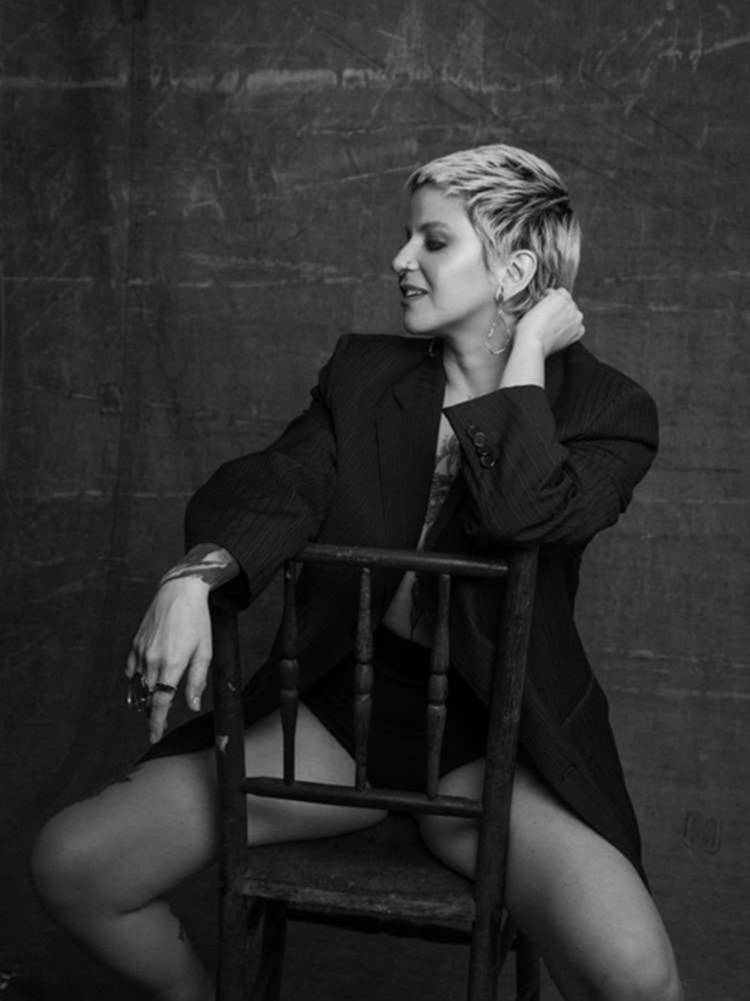 Mulher não-modelo com mais de 40 anos em ensaio fotográfico, usando vestido blazer preto + calcinha de cós alto, sentada de forma sensual em cadeira