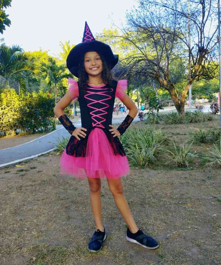 Garota com fantasia de bruxa infantil. Cores predominantes: preto e pink . Usa: vestido, luvas, tênis e chapéu pontudo