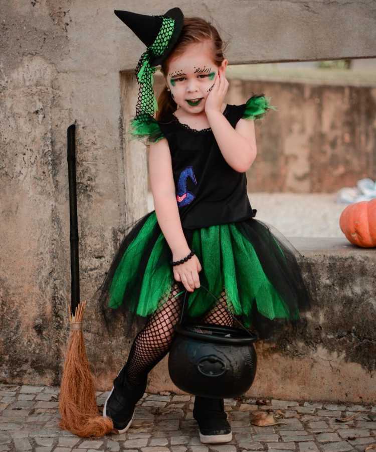 Garota com fantasia de bruxa infantil. Cores predominantes: preto e verde. Usa: Vestido, tiara de chapéu, meia calça arrastão, maquiagem, vassoura de brinquedo e balde em formato de caldeirão